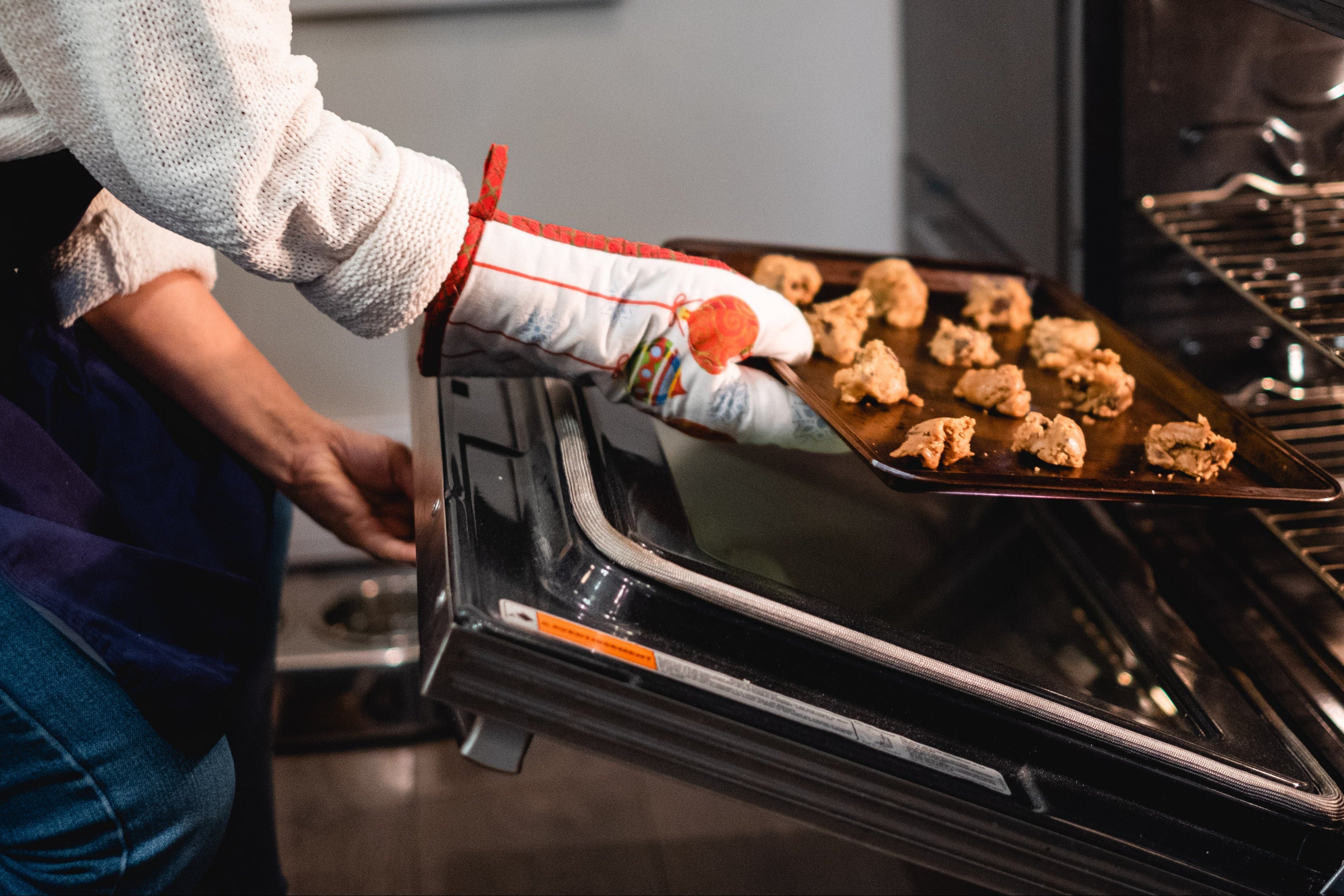 A person wearing an oven mitt pulls a baking tray of cookies out of a black built-in oven in a kitchen setting.